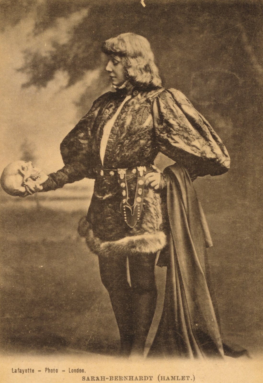 A sepia photograph of Sarah Bernhardt in costume as Hamlet, standing with one hand on her hip and holding out a skull in the other