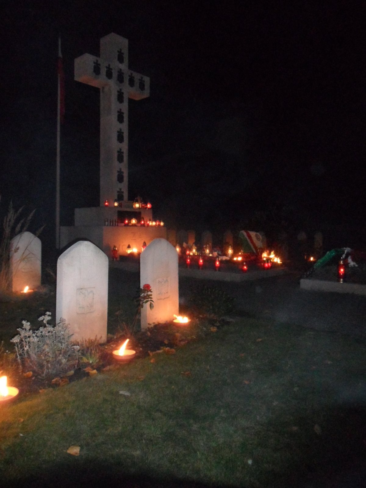 Candles glowing on the ground among white headstones at night, with a tall pale cross rising behind them in a cemetery