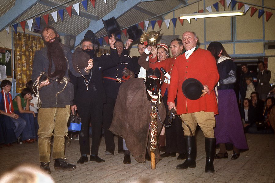 A group of costumed mummers in top hats, a red hunting coat and a long hooded cloak standing on a hall floor under bunting, with an audience looking on