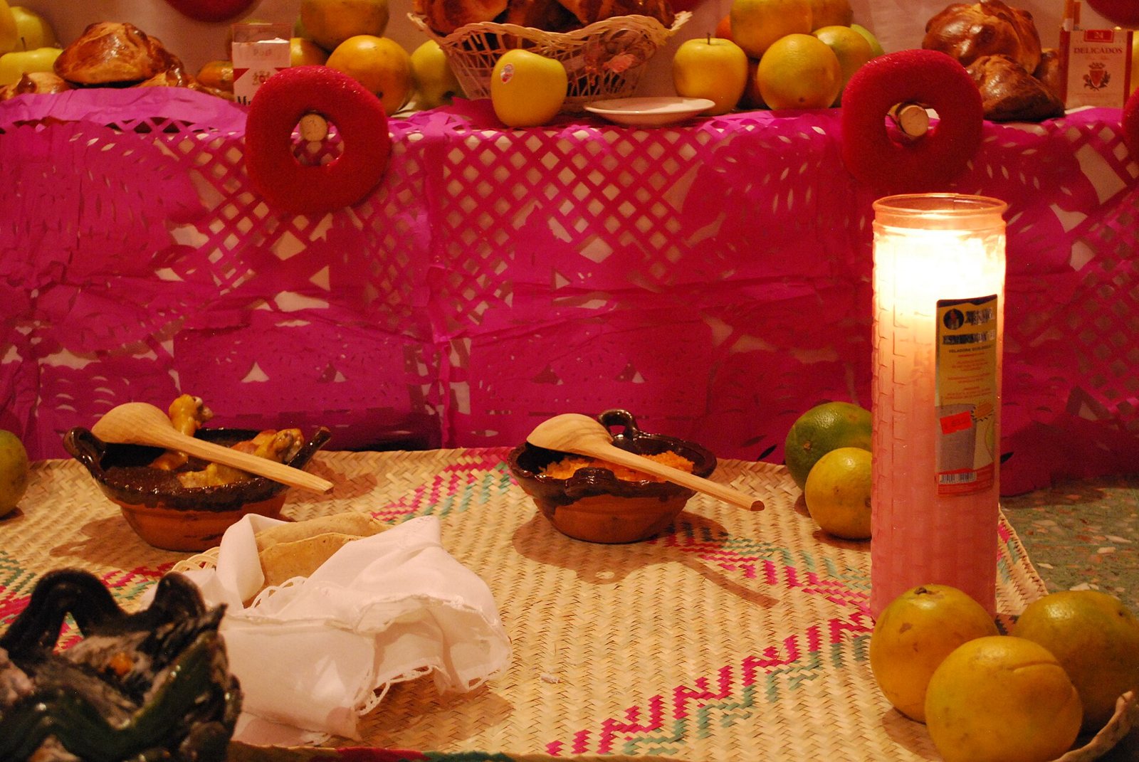 Una pequeña ofrenda casera sobre un petate bajo banderines rosas de papel picado: una vela alta encendida, cuencos con cucharas de madera, un paño blanco doblado y naranjas, con más fruta y pan en la repisa de arriba