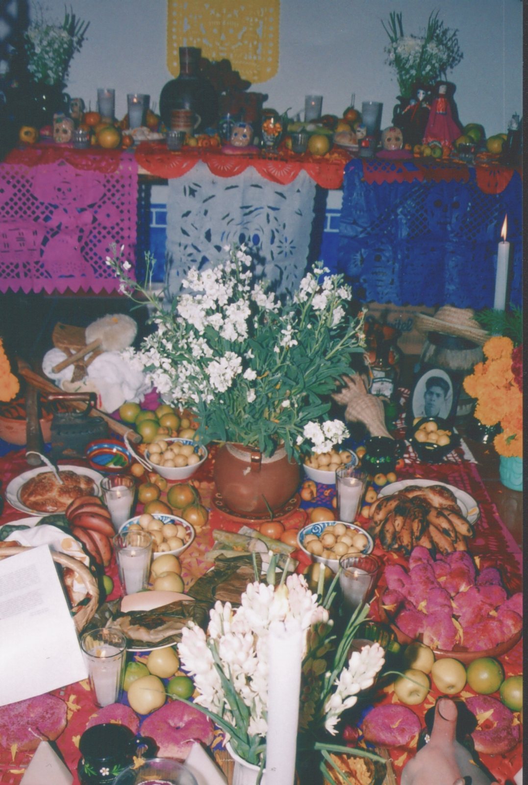 Una ofrenda casera con una mesa de fruta, pan y platillos bajo banderines de papel picado y flores blancas en un florero, con una segunda ofrenda sobre un petate en el suelo, más abajo