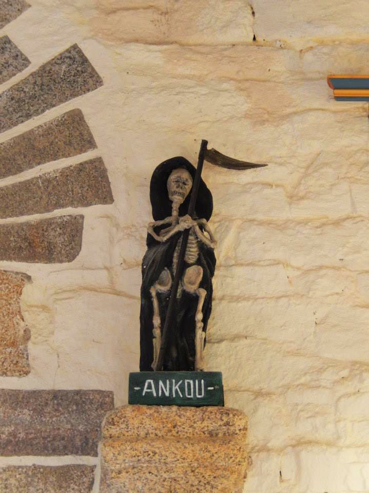 A small skeleton figure in a black hooded cloak holding a long scythe on a granite shelf, beside a green sign that reads Ankou