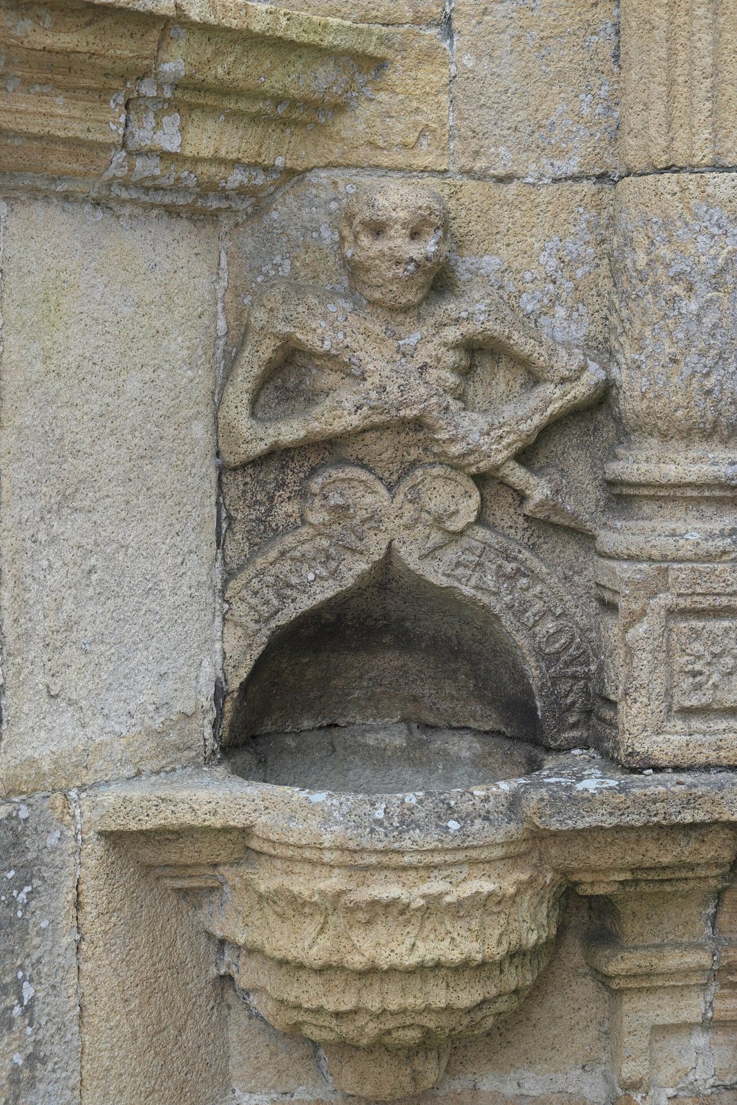 A carved stone skeleton with crossed arms and a dart, set in a niche above a stone holy-water basin on the ossuary wall
