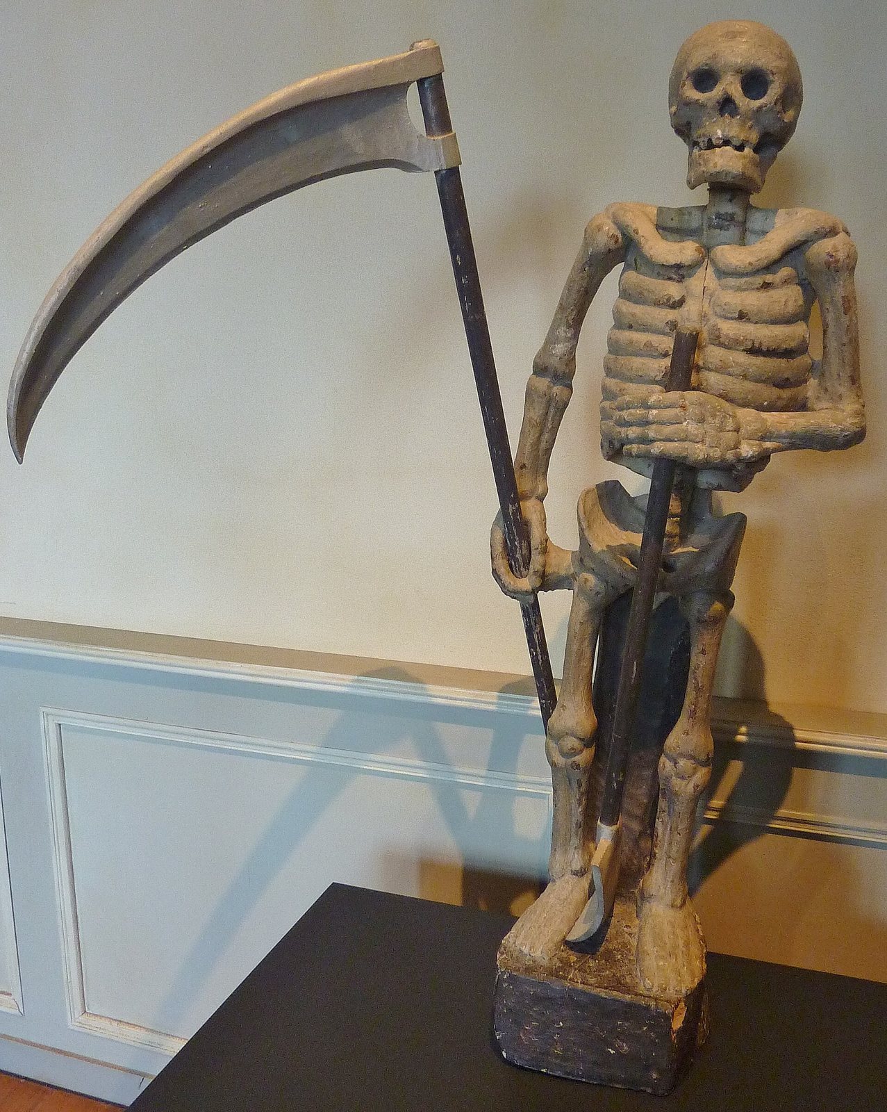 The wooden Ankou of Ploumilliau, a painted skeleton with a very large scythe and a shovel, standing on a black base against a pale wall