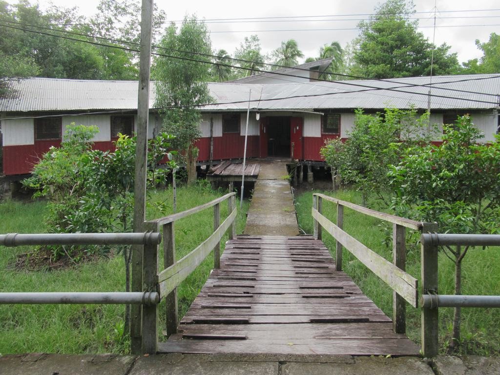 A long wooden walkway with rails leading across green grass to the entrance of a red and white building with a metal roof, surrounded by trees