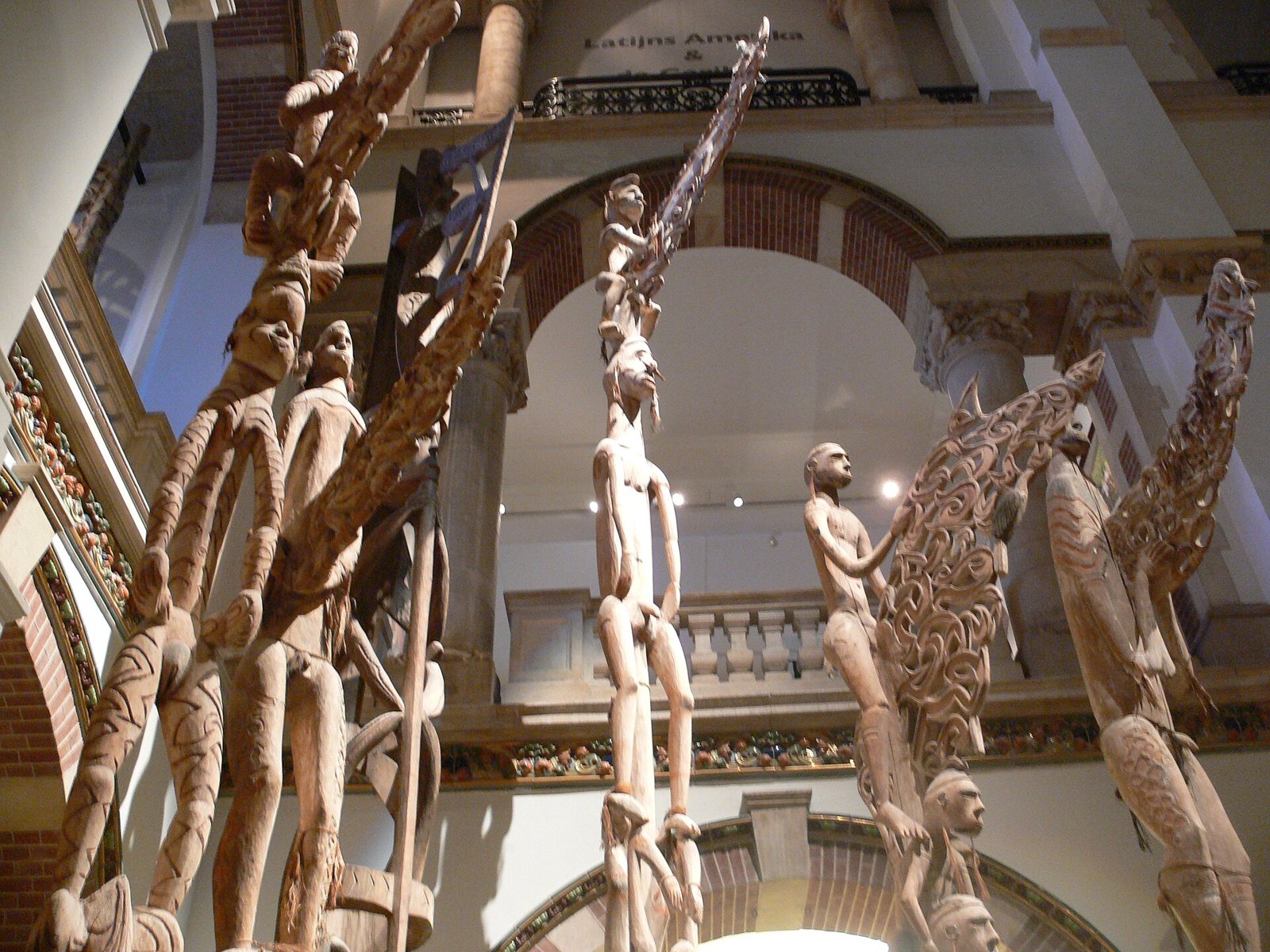 Several carved Asmat bisj poles seen from below in the central hall of the Tropenmuseum, their stacked figures and openwork tops rising toward arched galleries