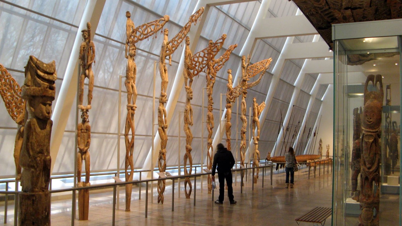 A row of tall carved wooden Asmat bis poles, each with stacked human figures and an openwork flag-like projection at the top, standing along a museum gallery with a slanted glass wall while visitors look on