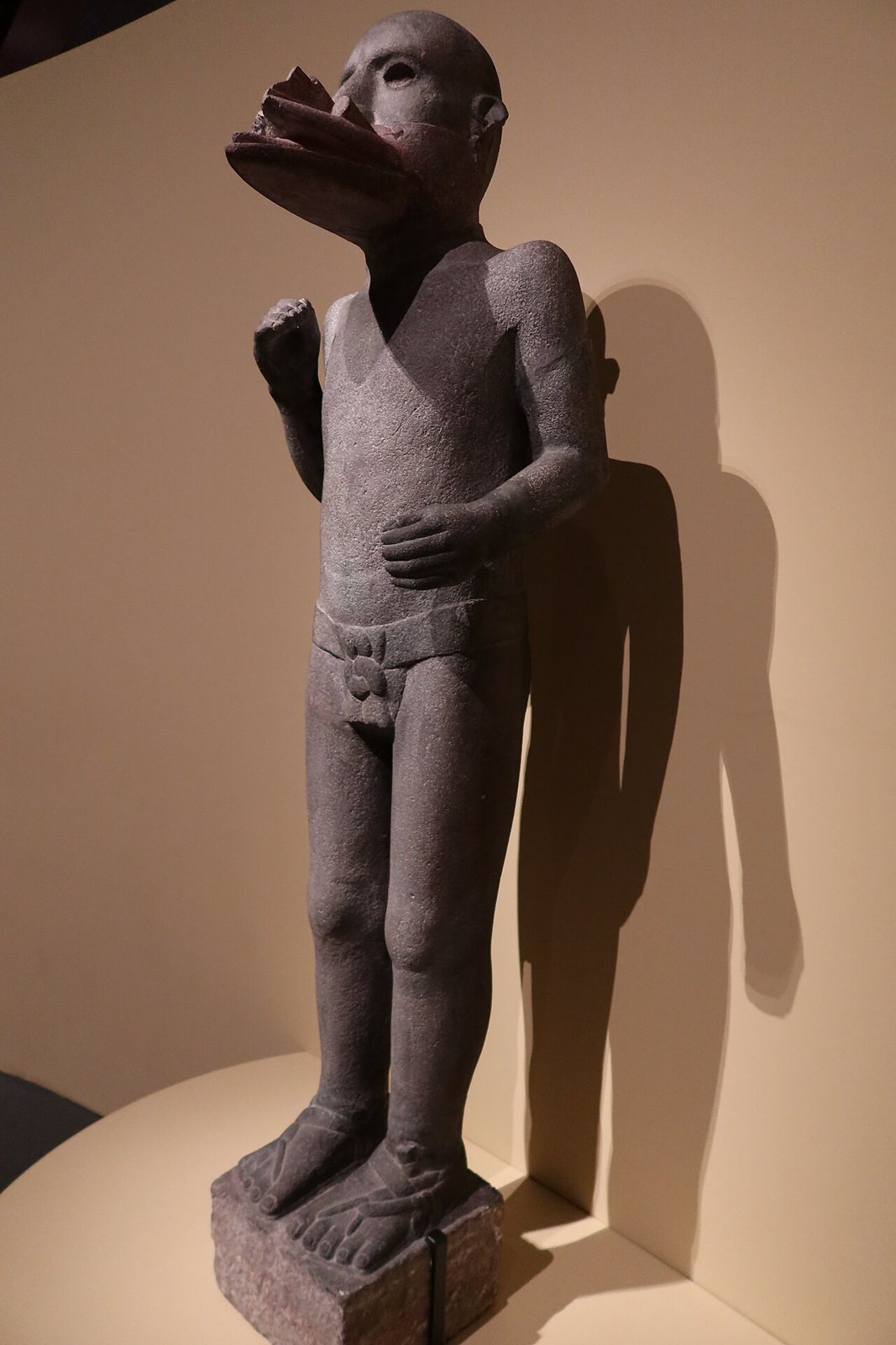 A standing basalt figure of the wind god Ehecatl wearing a loincloth and sandals, his face covered by a projecting beak-like mask, displayed in a museum