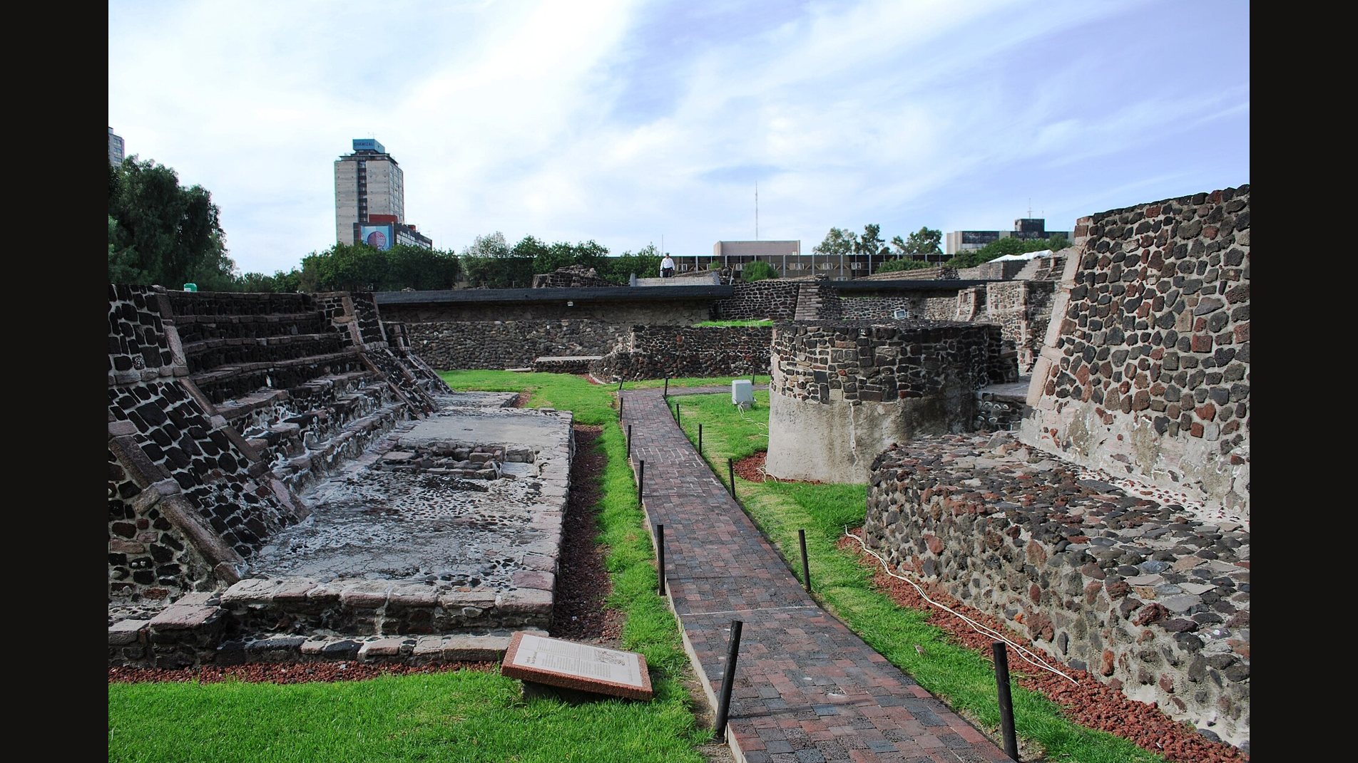 The Tlatelolco archaeological zone in Mexico City: a brick path runs between dark volcanic stone platforms and stepped temple bases on green lawns, with modern towers behind
