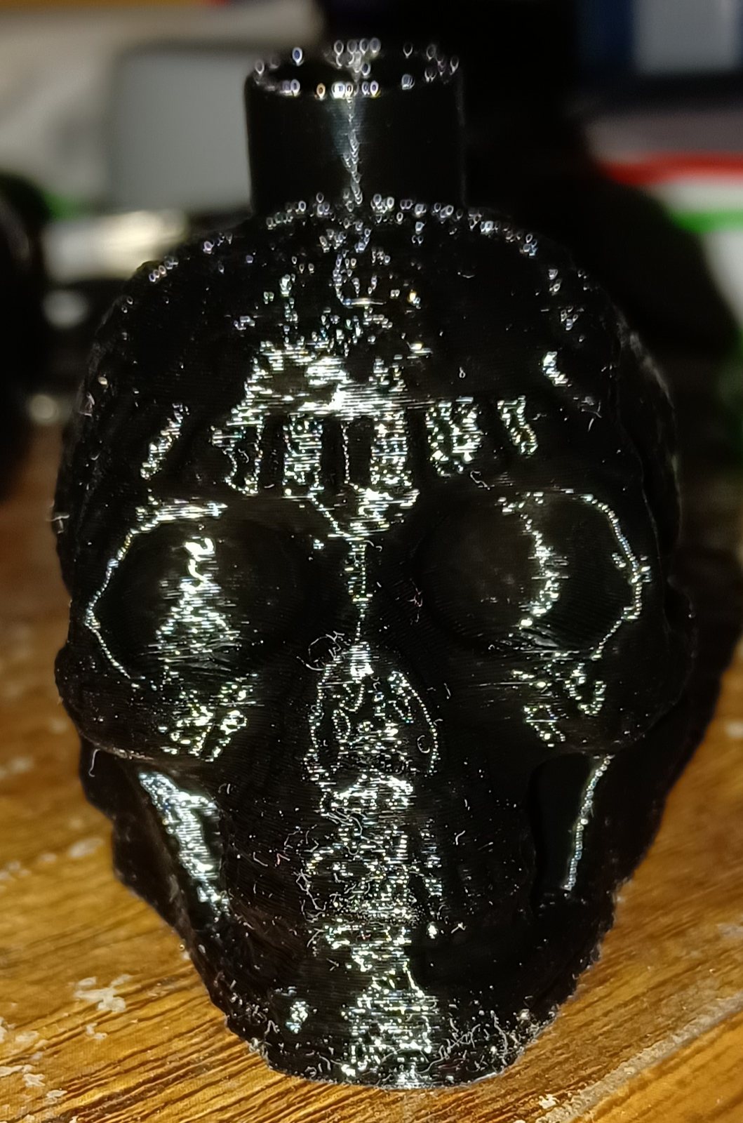 A glossy black 3D-printed skull whistle standing on a wooden table, with a short mouthpiece on top of the skull and visible print lines across the face