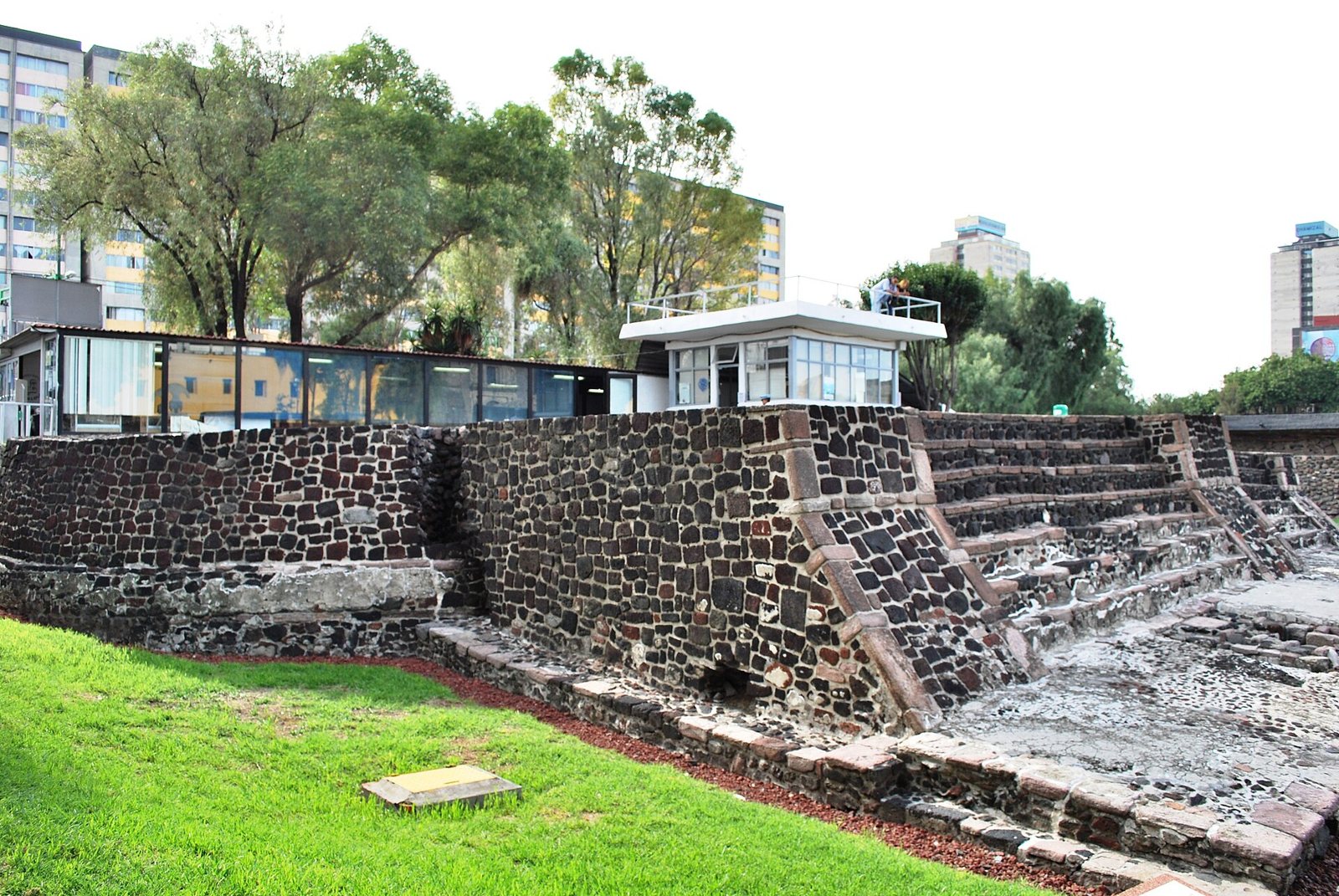 The stepped base of the Temple of Ehecatl-Quetzalcoatl at Tlatelolco, built of dark stone blocks, with a lawn in front and apartment buildings and trees behind