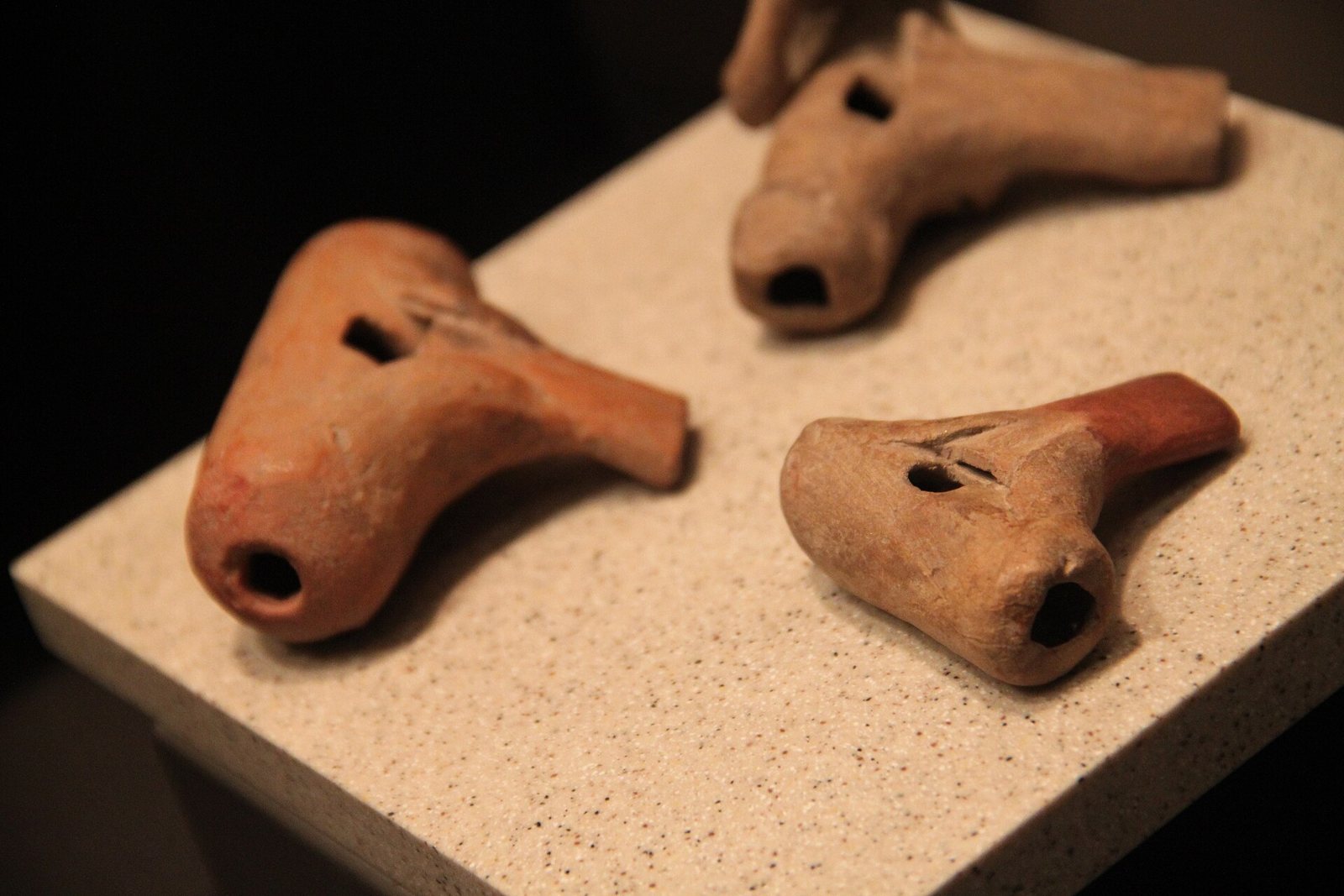 Three small clay whistles in reddish and buff tones resting on a speckled stone display block, each with a round mouth opening and a rectangular sound hole