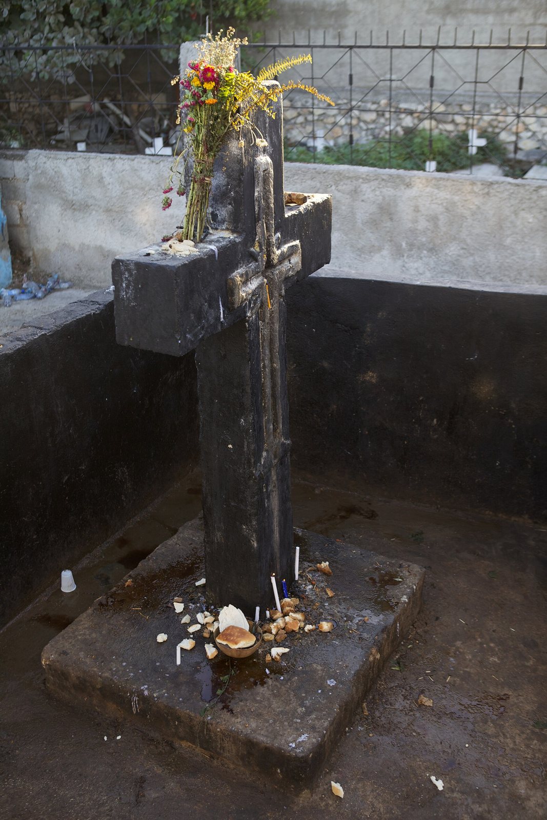 A tall black cross on a square stone base inside a walled tomb enclosure, with dried flowers tied to the top and small offerings at its foot