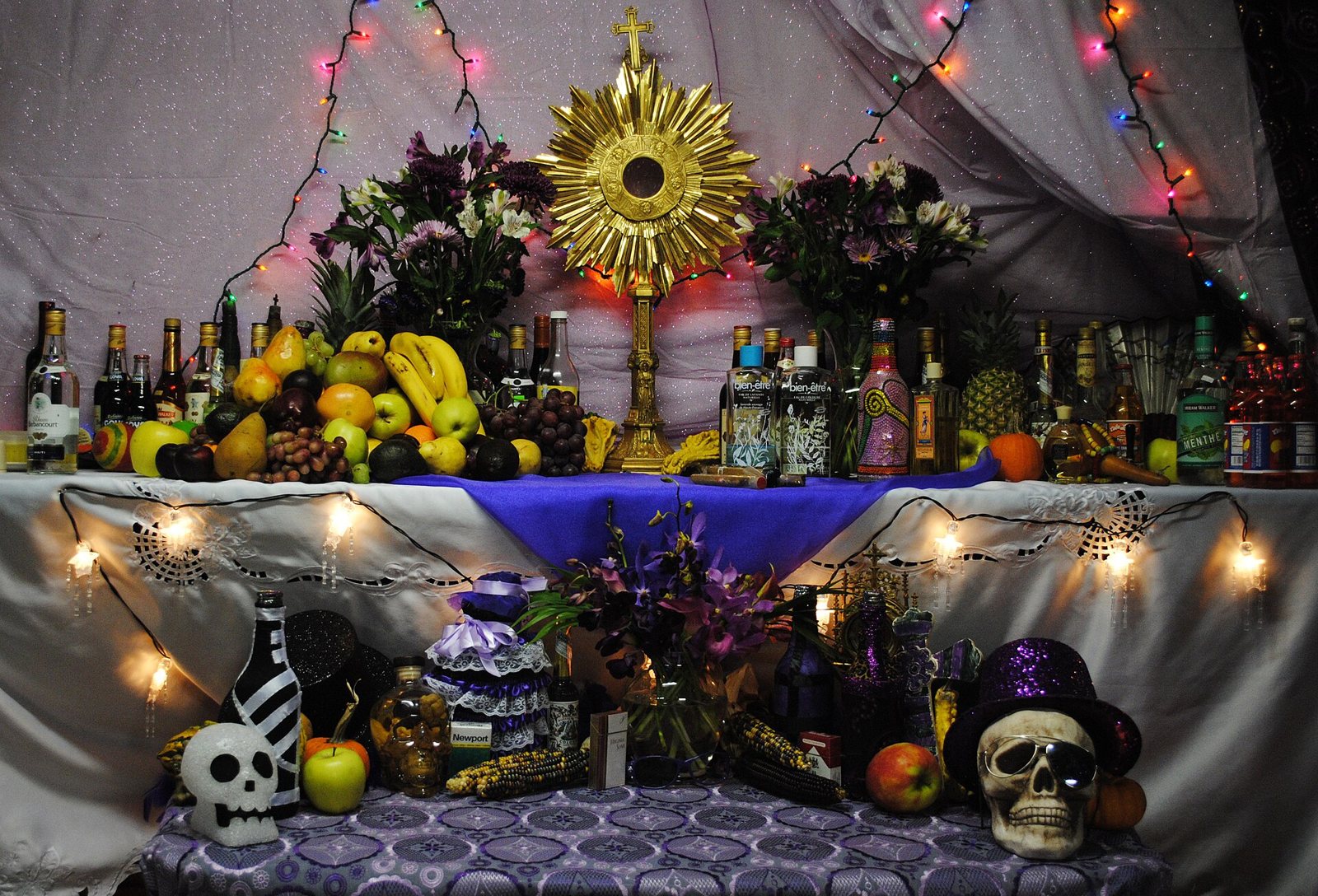 A Vodou altar hung with string lights, crowded with fruit, flowers and bottles under a gold sunburst, with two skulls, one in a purple top hat, along the bottom tier