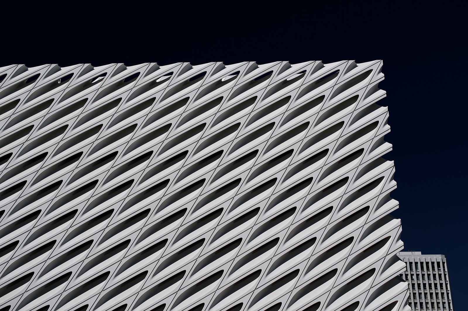 The honeycomb-patterned white exterior of The Broad museum in Los Angeles seen from below against a deep blue sky, with a tower block behind its corner