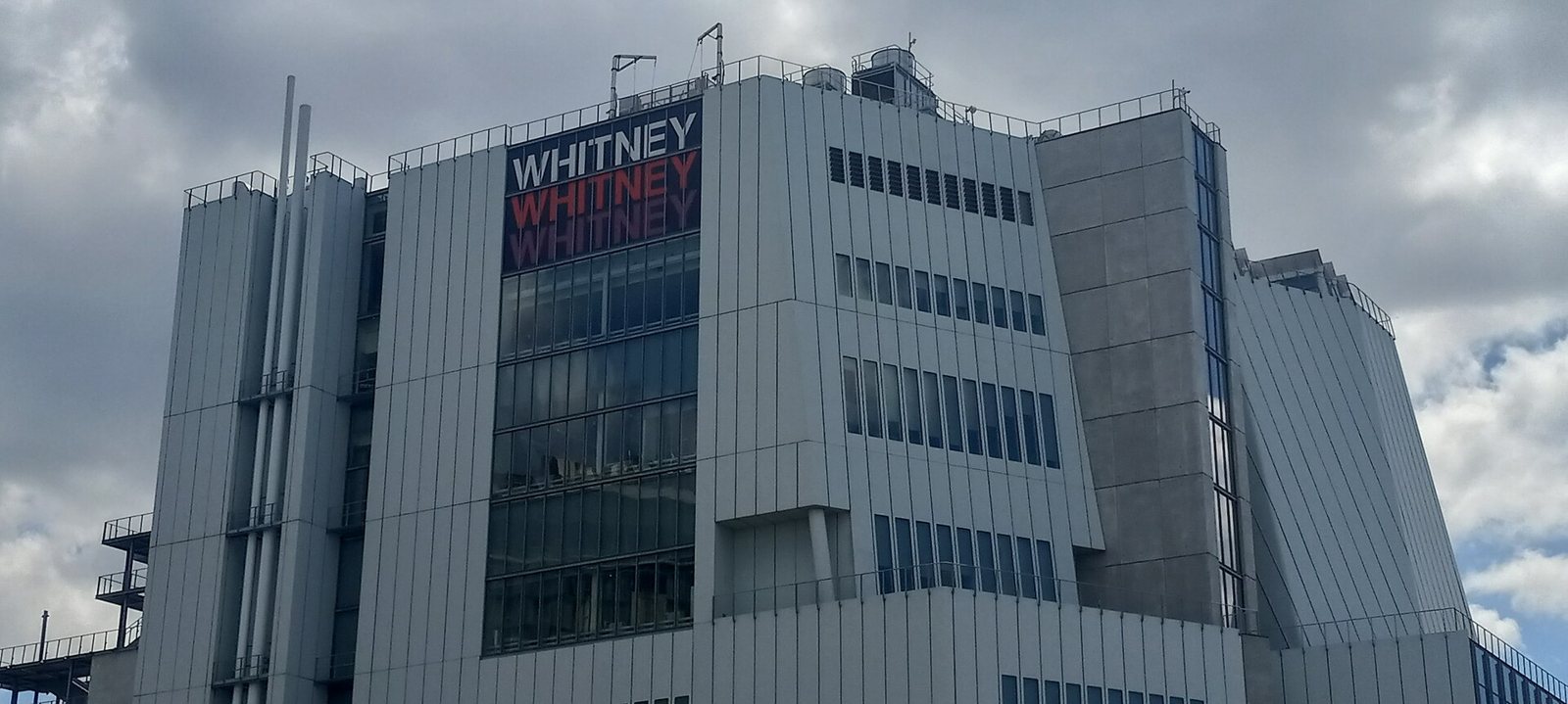 The gray, angular Whitney Museum building in New York under a cloudy sky, with the museum name on a banner near the top