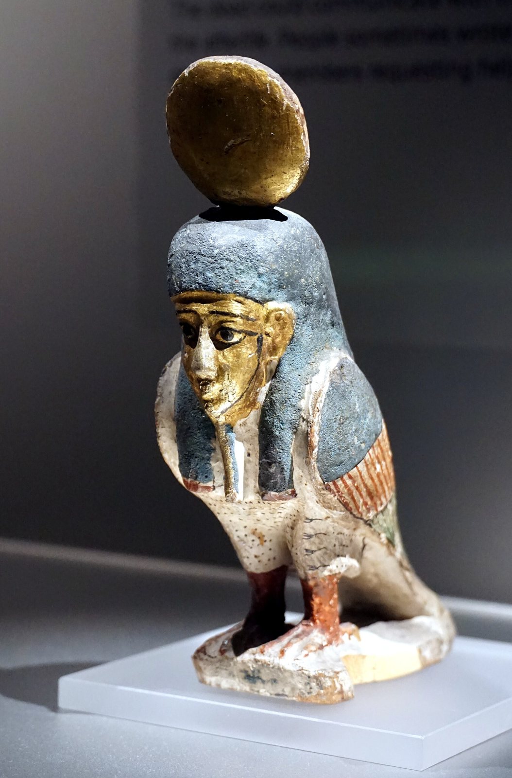 A painted wooden figure of a bird with a human head, blue headdress and a gold disc on top, standing on a clear stand in a museum case