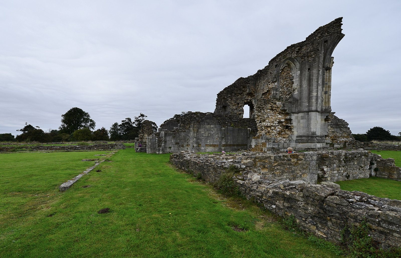 The ruined stone walls of Thornton Abbey in Lincolnshire rising from a mown green lawn under a grey sky, with trees on the horizon