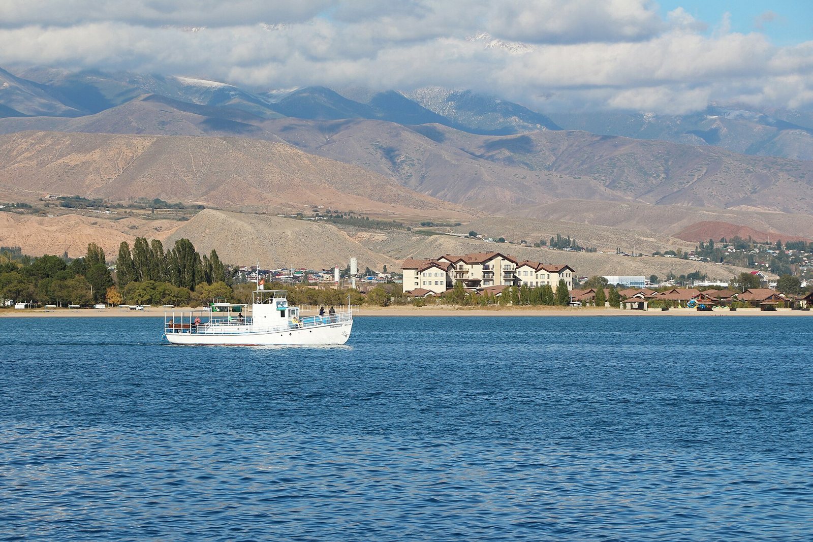 Lake Issyk-Kul in Kyrgyzstan, with a small boat on blue water, a town on the shore and dry brown mountains behind