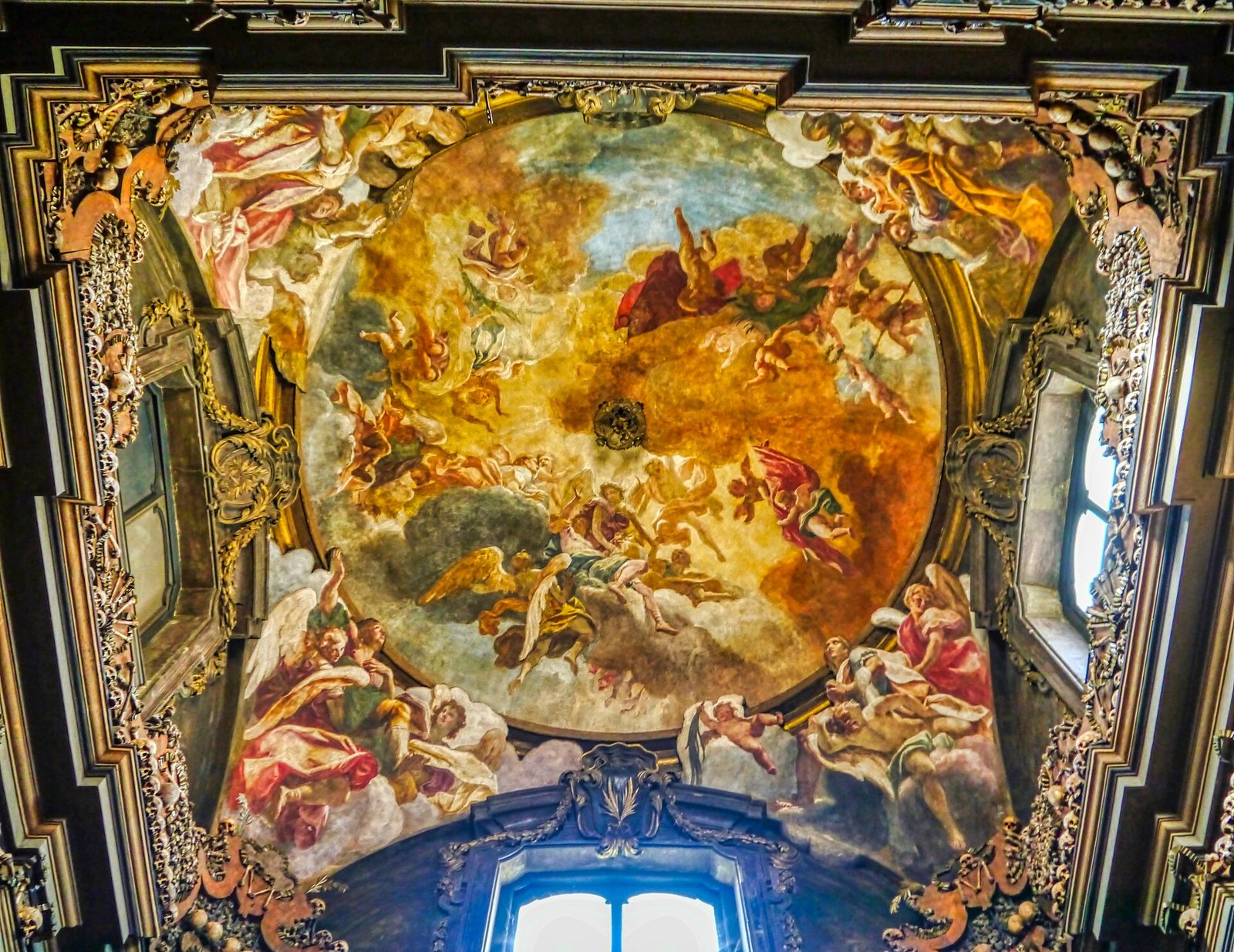 The painted ceiling of the ossuary: a round fresco of angels and figures among golden clouds and blue sky, inside an ornate gilded frame with a window below