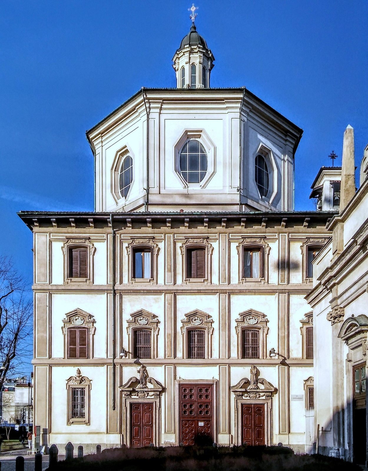 The cream facade of San Bernardino alle Ossa in Milan, with rows of arched windows, three red doors and an octagonal lantern on top, under a deep blue sky