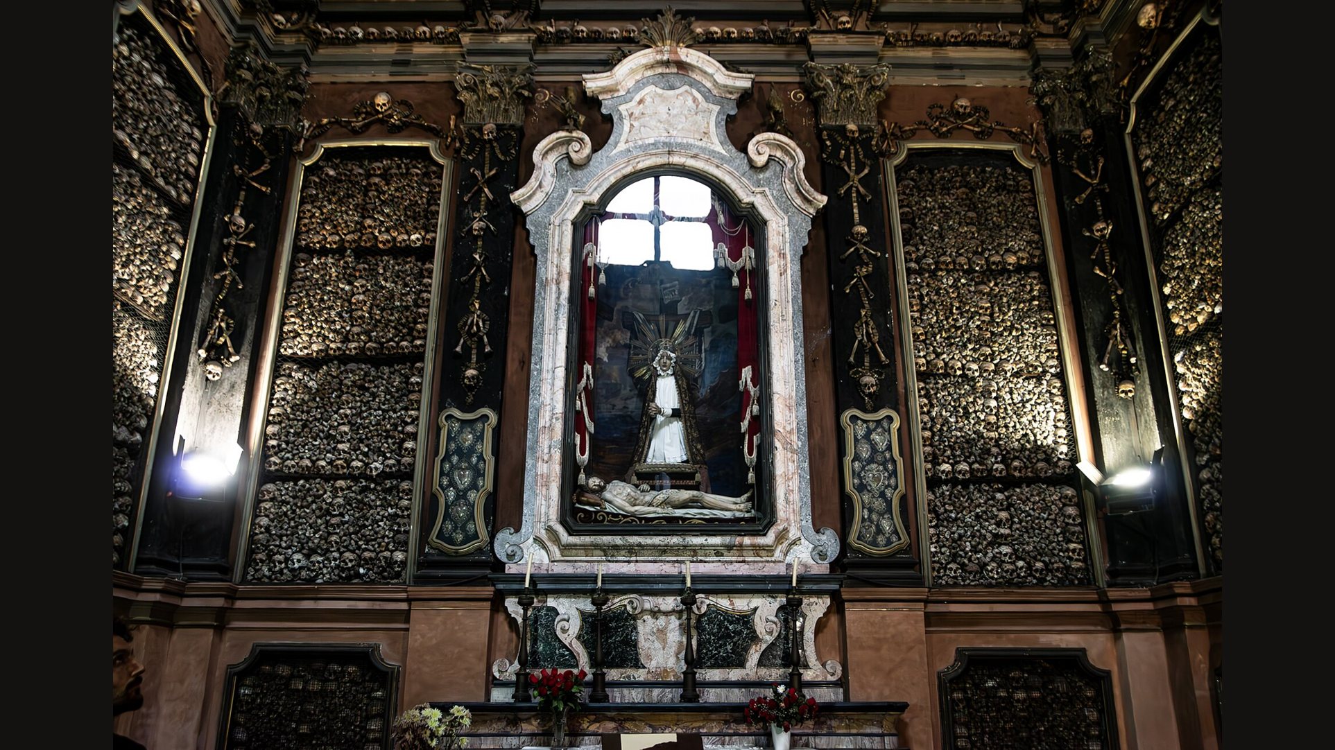 The altar of the ossuary of San Bernardino alle Ossa in Milan, a white marble frame holding a small figure, set into walls covered in skulls and bones and lit from the sides