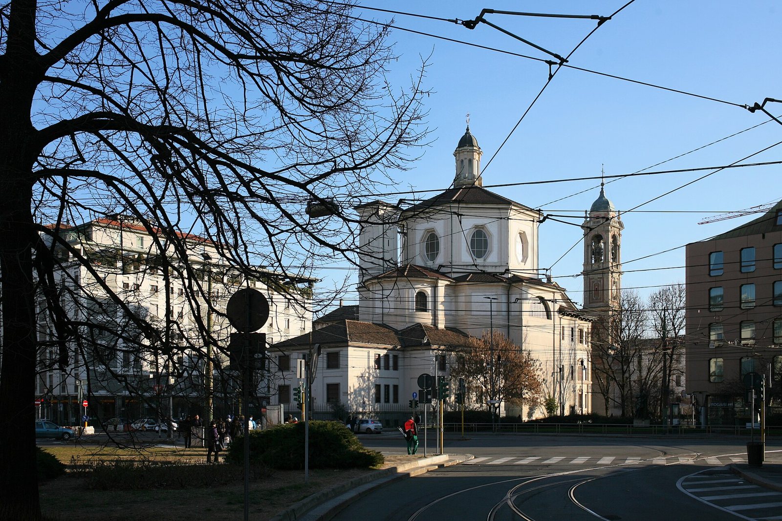 The San Bernardino and Santo Stefano complex seen across a Milan street, a pale domed church with a bell tower, framed by a bare tree and tram wires