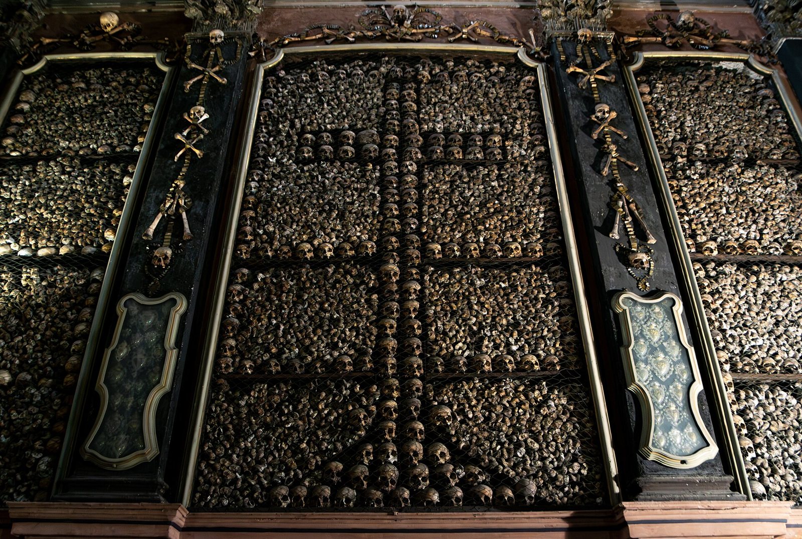 A wall of the ossuary with thousands of skulls and bones set into dark panels, with a cross picked out in skulls at the centre between gilded frames