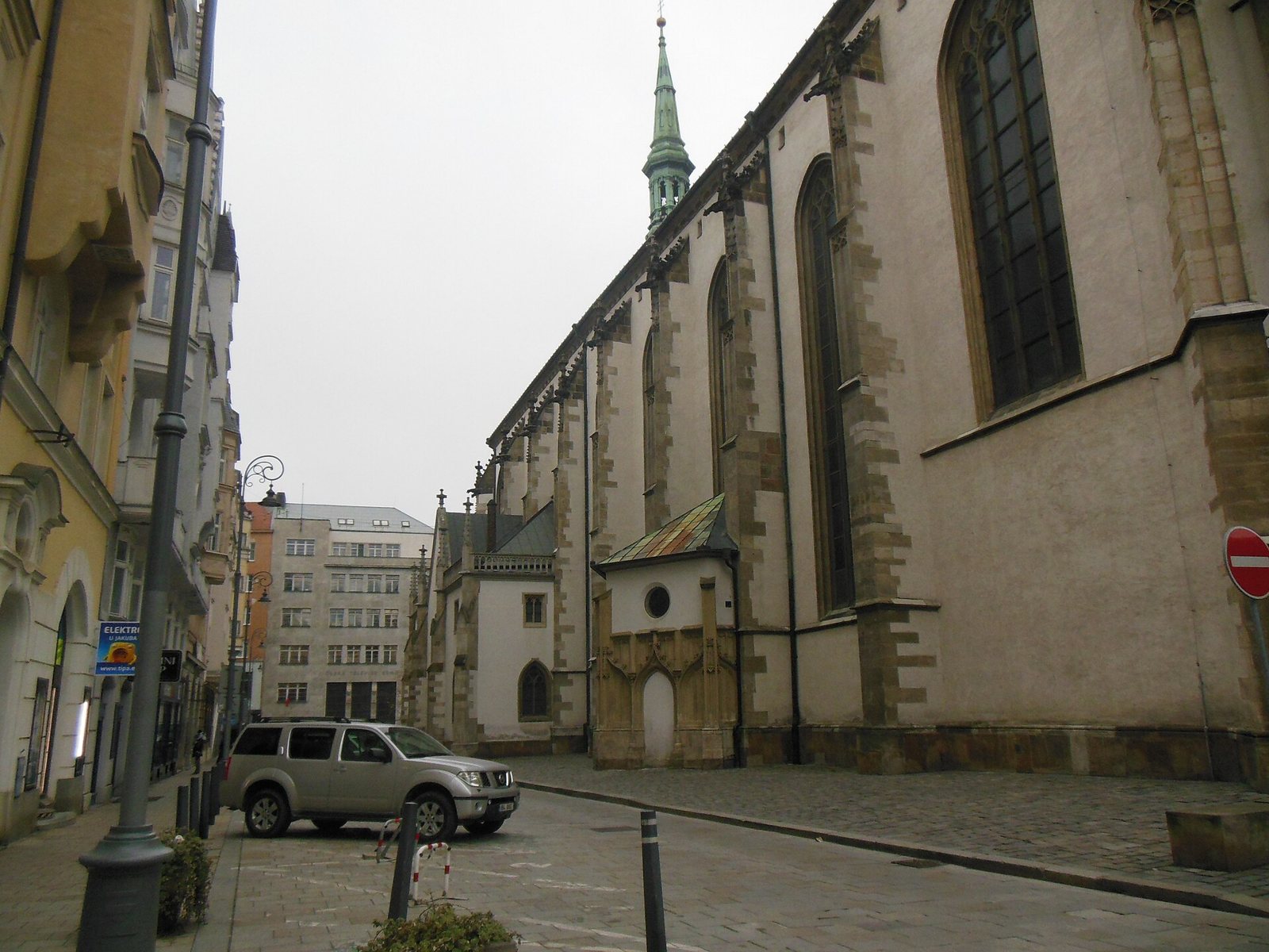 The long stone side of St James's Church in Brno seen from the narrow square, with its tall pointed windows and green spire
