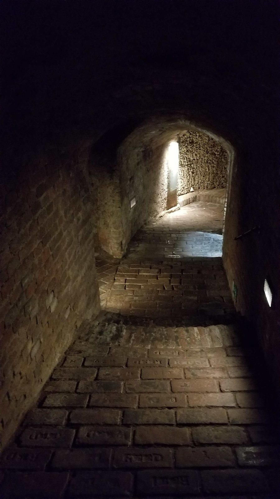 A narrow brick-vaulted passage in the Brno ossuary with a stone floor, lit by a single light at the far end