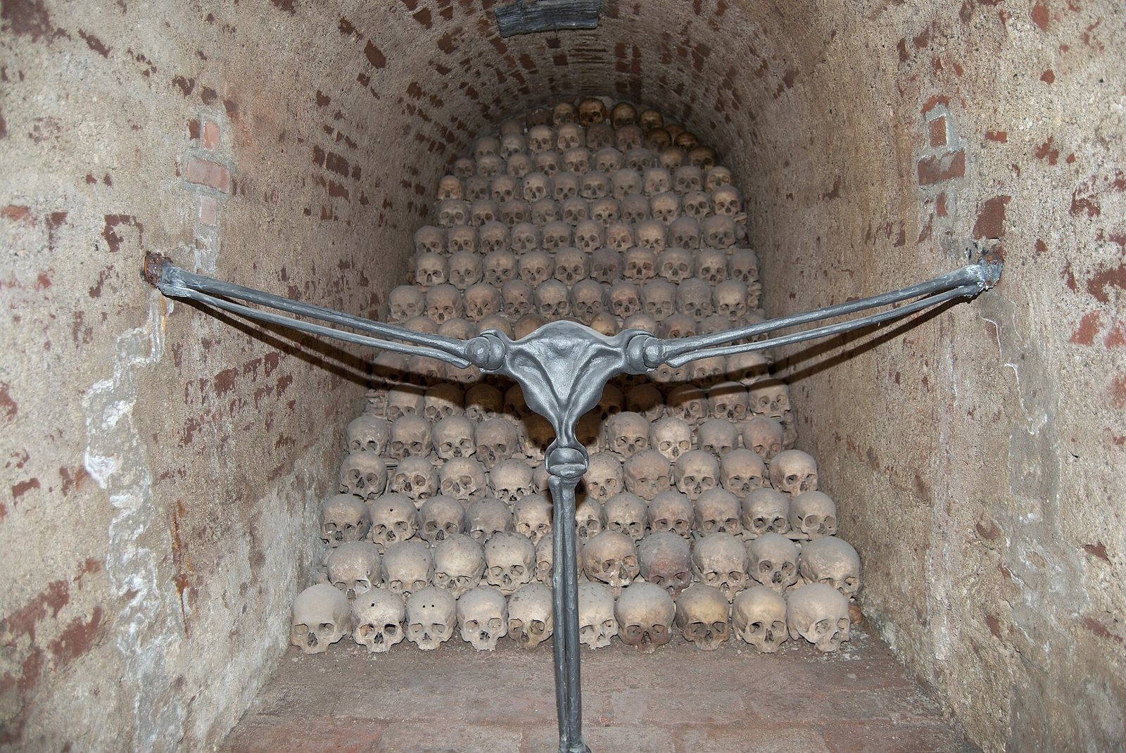 A pyramid of skulls in a brick niche of the Brno ossuary, with a modern wrought-iron barrier shaped like outstretched arms across the front