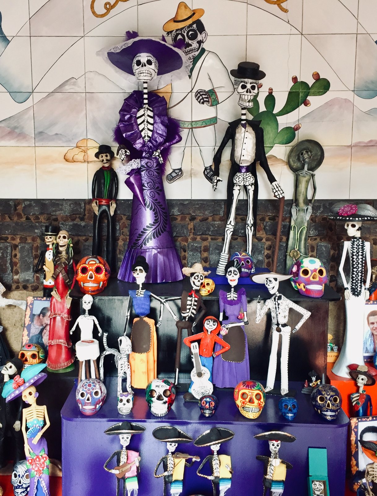Repisas con figuras de esqueletos pintados con vestidos, trajes y sombreros, con calaveras de azúcar y una pared de azulejos detrás, montadas como ofrenda de Día de Muertos