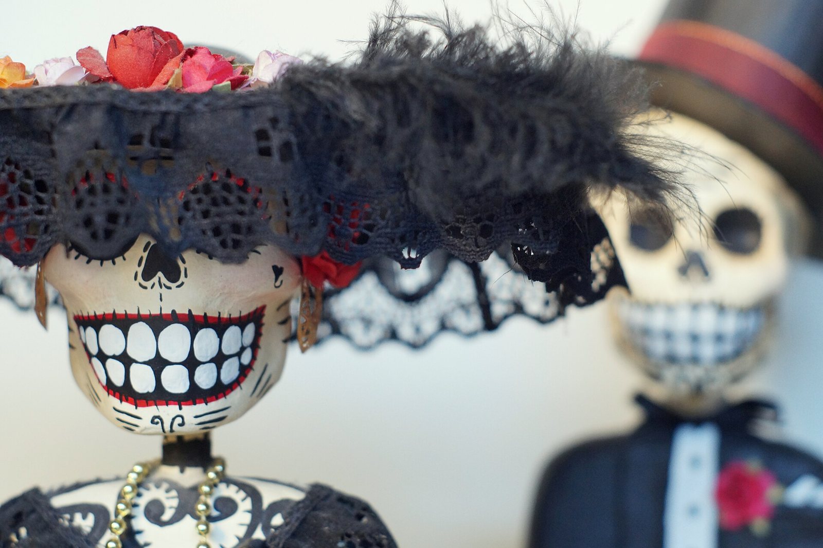 Vista cercana de dos figuras de cartonería: una Catrina sonriente con flores y un sombrero de encaje negro y plumas, y detrás, desenfocado, un Catrín con sombrero de copa y traje oscuro