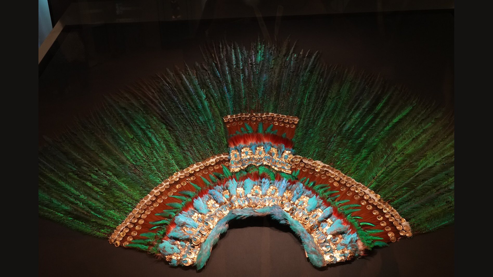 A fan-shaped crown of long green quetzal feathers with a band of red, blue and gold ornaments, displayed in a dark museum case