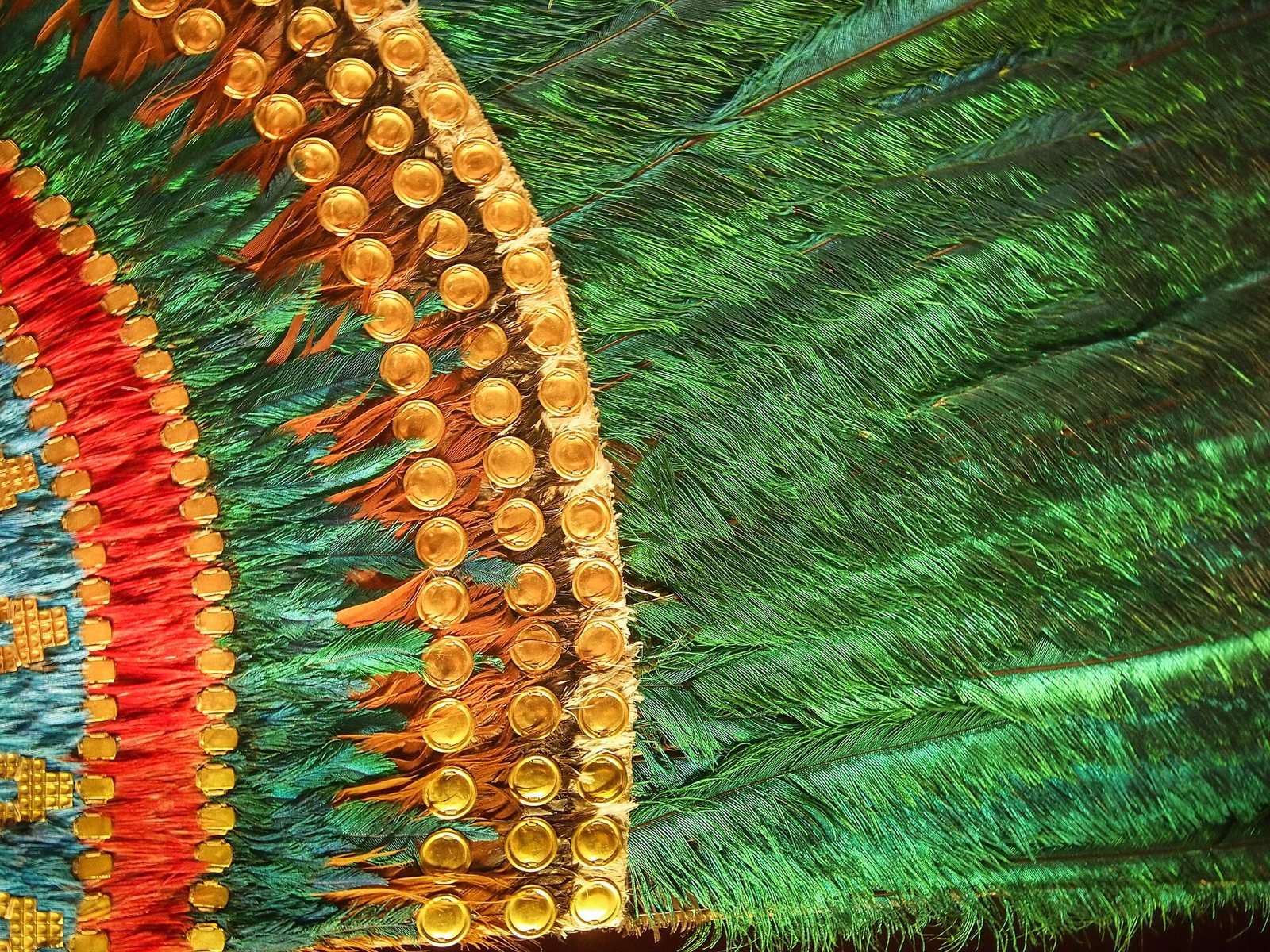 Close-up of green quetzal feathers and rows of gold discs on a red, turquoise and orange band
