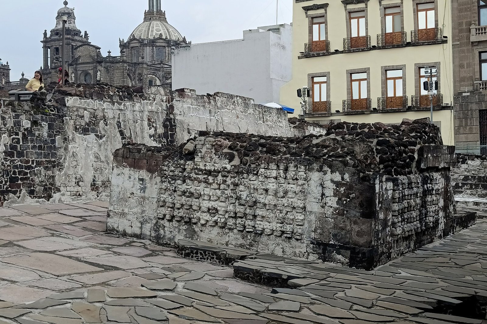 Stone ruins of the skull rack platform at the Templo Mayor, its wall carved with rows of skulls, with a colonial cathedral dome and buildings behind