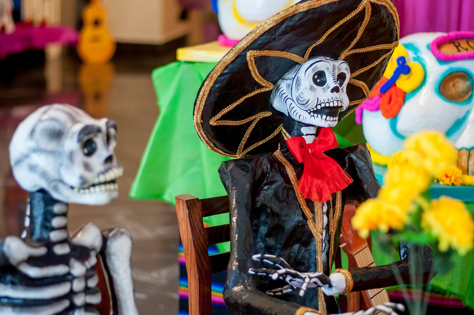 Papier-mâché skeletons on a Día de Muertos display: one in a black charro outfit and wide sombrero with a red bow, seated beside yellow marigolds, and another skeleton at left