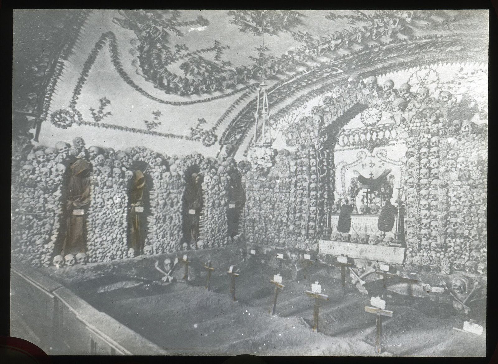 Diapositiva de vidrio en blanco y negro de la Cripta de los Capuchinos, con ornamentos de huesos en la bóveda y los muros y cruces de madera en el suelo