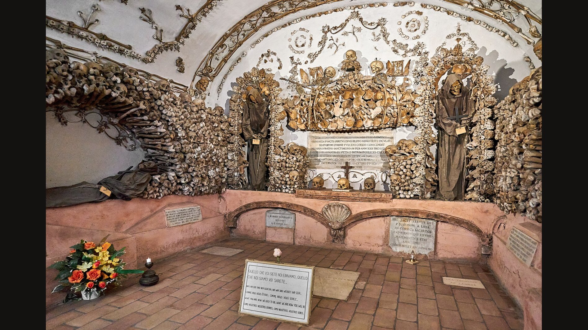 Una capilla de la Cripta de los Capuchinos en Roma con bóveda de ornamentos de huesos y esqueletos de frailes con hábito en nichos, sobre un suelo de ladrillo con un ramo de flores