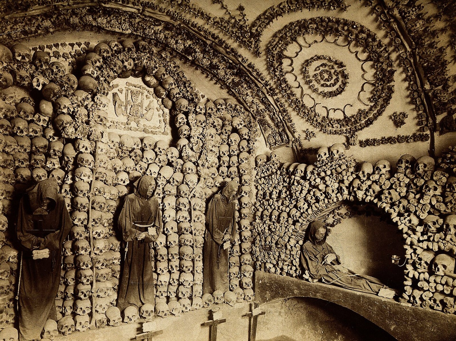 Fotografía en sepia de la Cripta de los Capuchinos con arcos y rosetones de huesos en los muros y tres restos de frailes con hábito en nichos