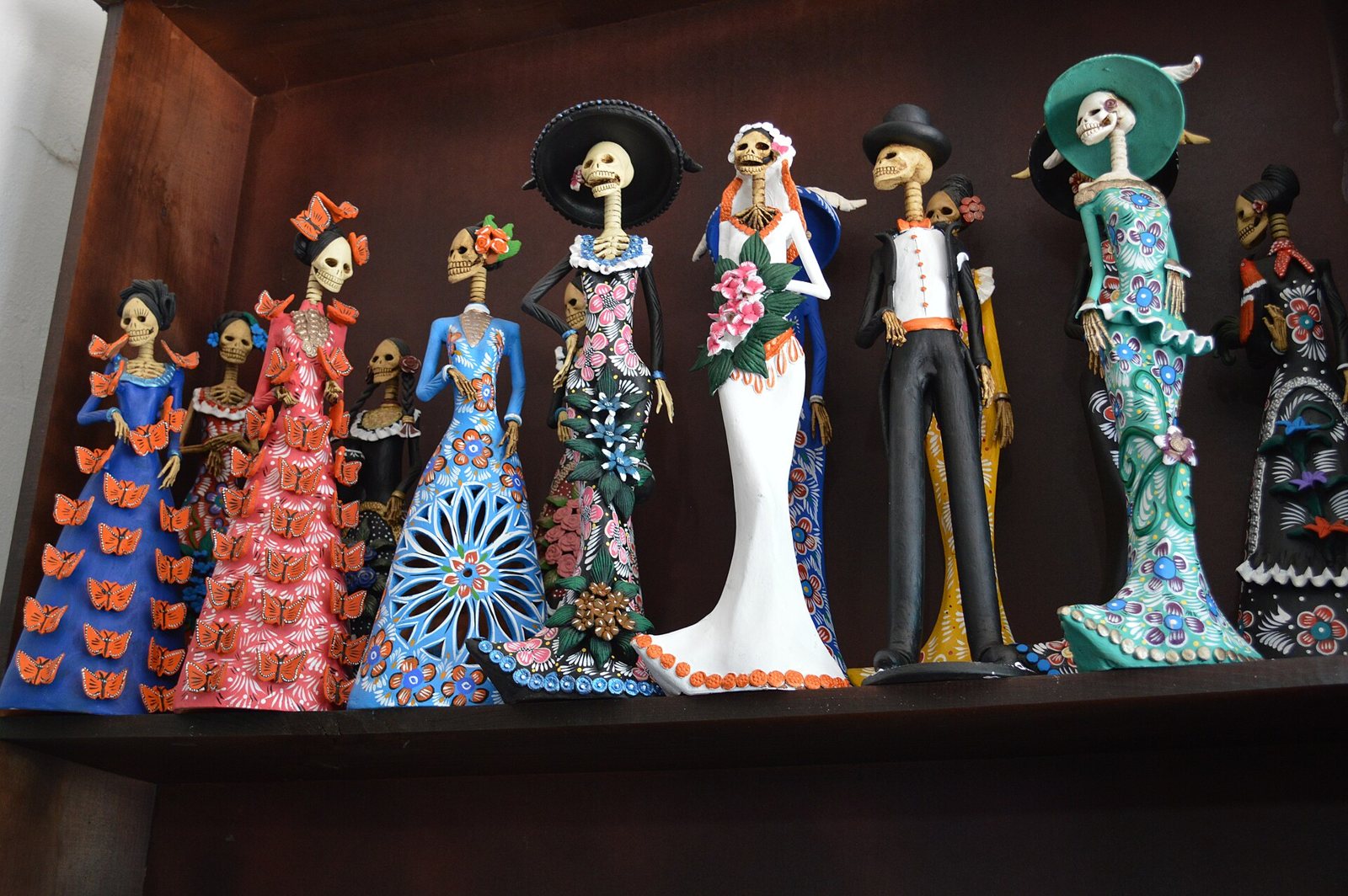 Seven painted clay Catrina and Catrín figures standing on a dark wooden shelf in a Capula workshop, in dresses and suits decorated with flowers and butterflies