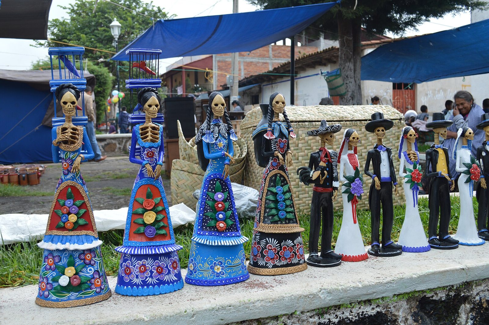 Blue-dressed clay Catrinas carrying cages on their heads and several black-suited figures lined up on a low wall in the main square of Capula, under blue awnings