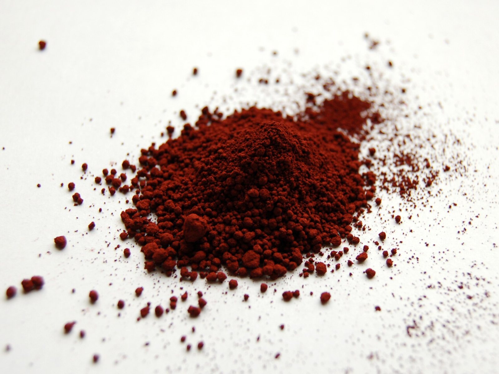 A heap of dark red-brown iron oxide powder on a white surface, with a scatter of loose grains around it