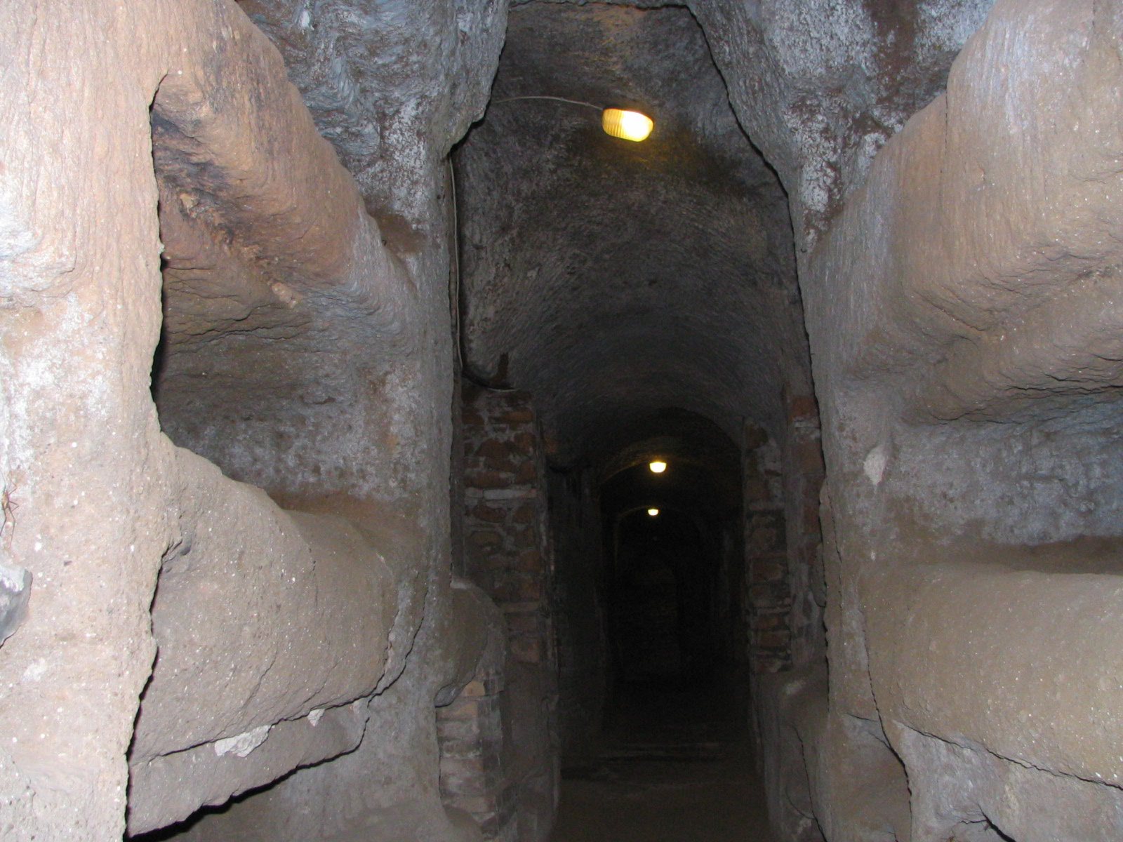 A narrow underground passage cut through brown rock, with burial recesses in the walls and small lamps lighting the way ahead