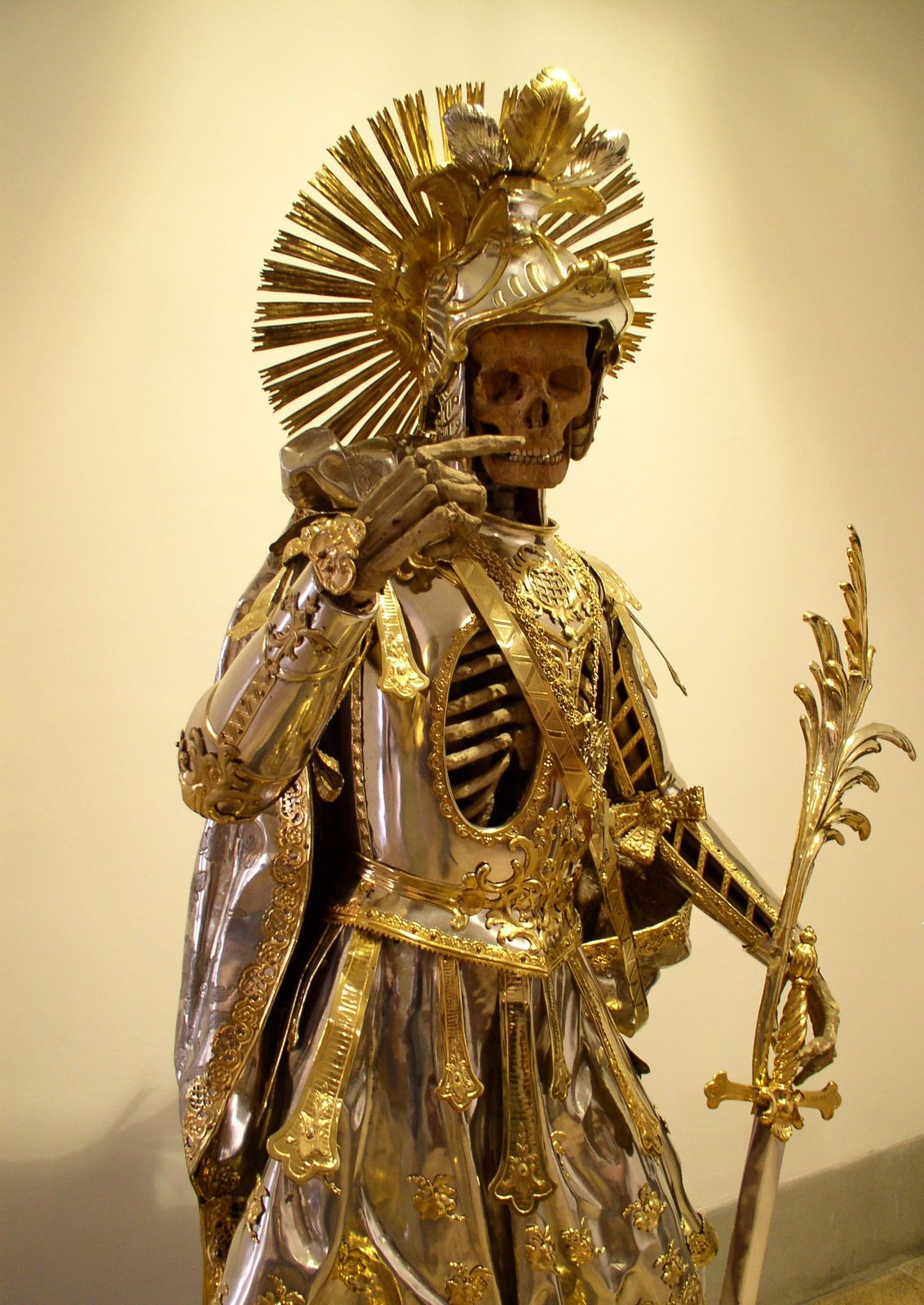 A standing skeleton dressed in silver and gilded armor with a sunburst helmet, one hand raised to the jaw and the other holding a tall gold martyr's palm