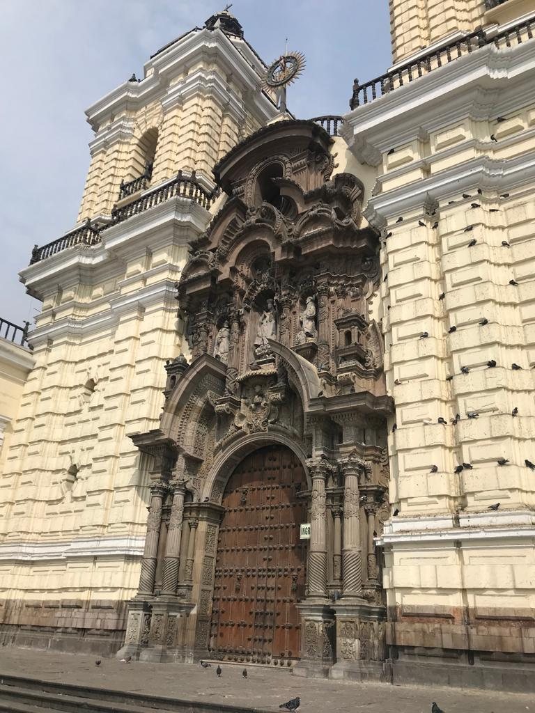 La fachada de piedra color crema de la iglesia de San Francisco en Lima, con una portada barroca tallada, altas puertas de madera y dos torres encima