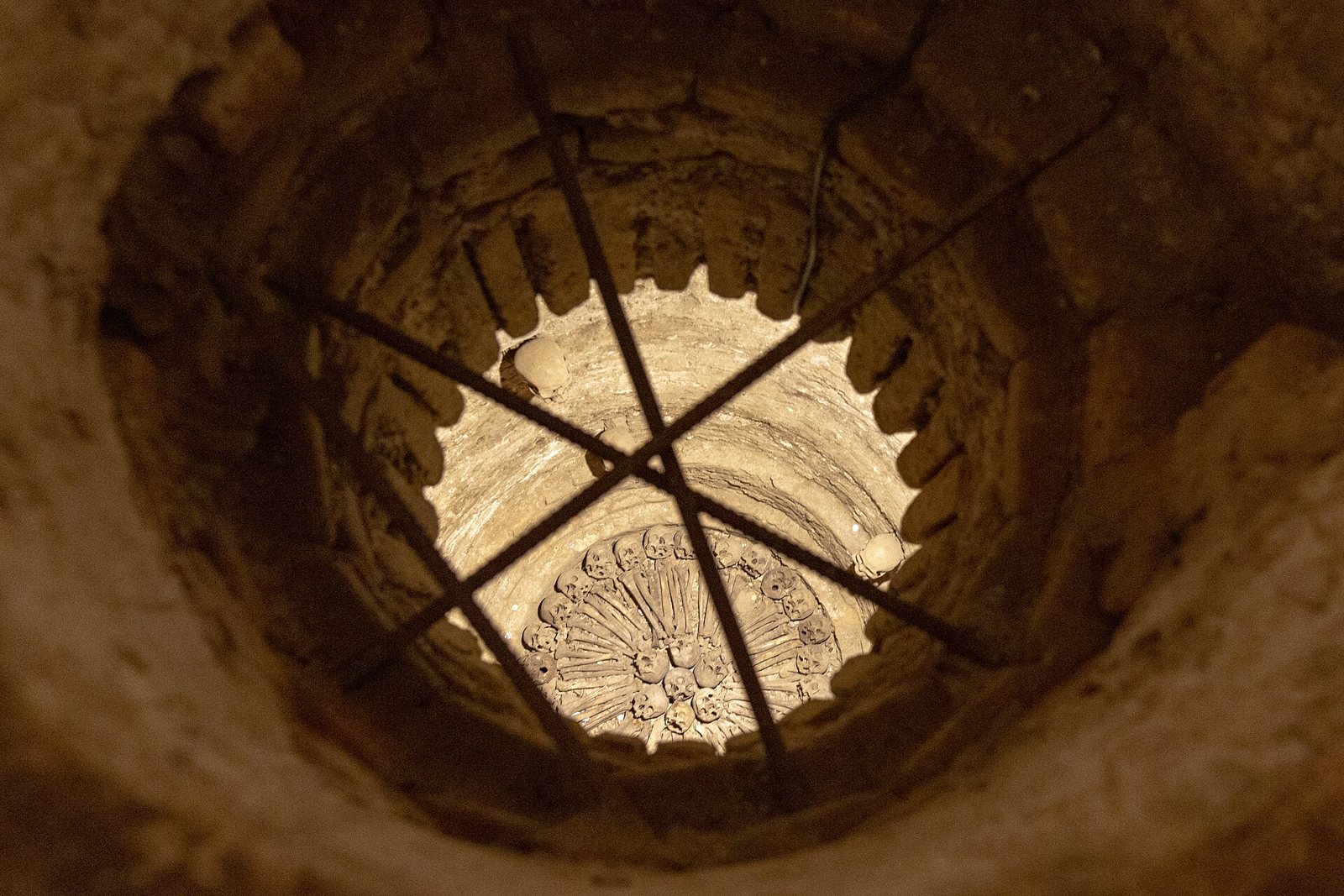 Vista hacia abajo de un pozo circular revestido de ladrillo en las catacumbas, atravesado por barras de hierro, con una rosa de piedra tallada en el fondo