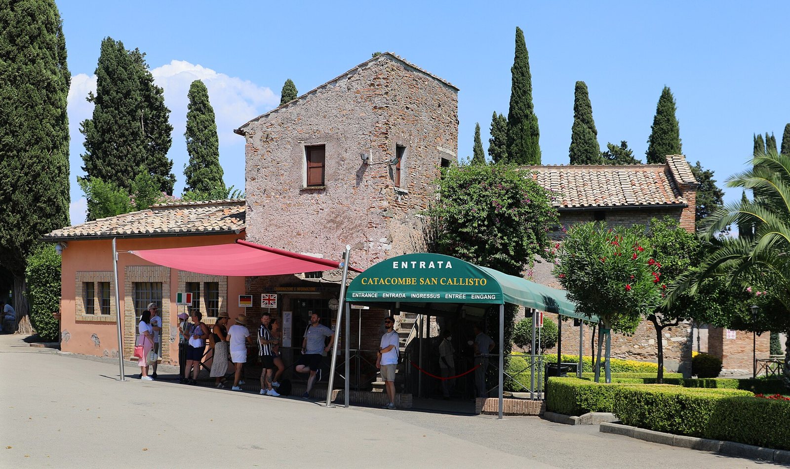 La entrada para visitantes de las catacumbas de San Calixto en un día soleado: un edificio bajo de ladrillo con un toldo verde que dice Entrata, cipreses altos detrás y un pequeño grupo de personas esperando