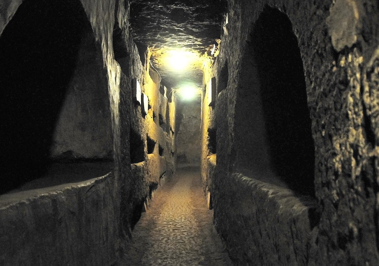 Una galería subterránea estrecha y de poca luz en las catacumbas de Domitila, con filas de nichos funerarios excavados en las paredes de roca a ambos lados y lámparas en el techo