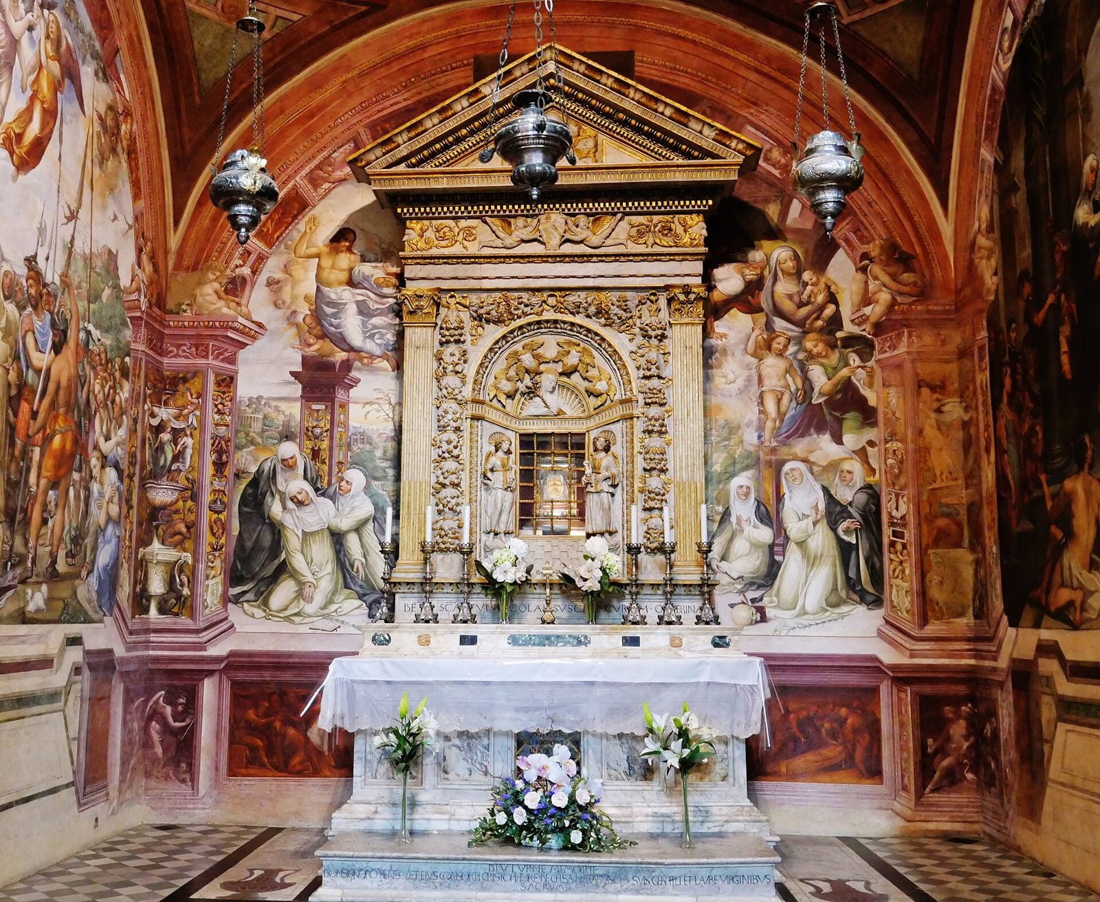The Chapel of Saint Catherine in San Domenico, Siena: a gilded marble altar frame in the centre, a white-draped altar with lilies, hanging lamps, and walls covered in colourful frescoes